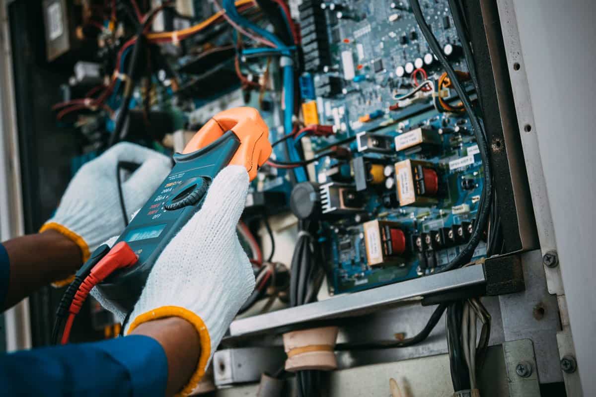 Technician testing electrical components on a control board using a clamp meter for safety compliance.